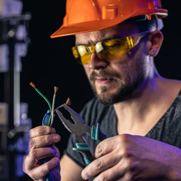 A male electrician in a protective helmet works in a switchboard with an electrical connecting cable.